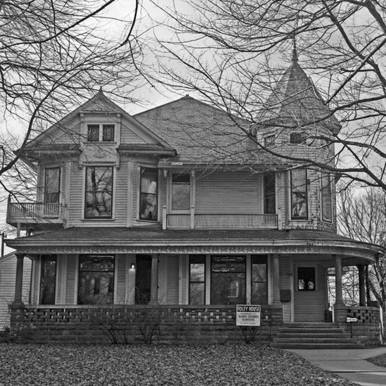 Black and white image of the Foley House, a large, Victorian style house with a wraparound porch.