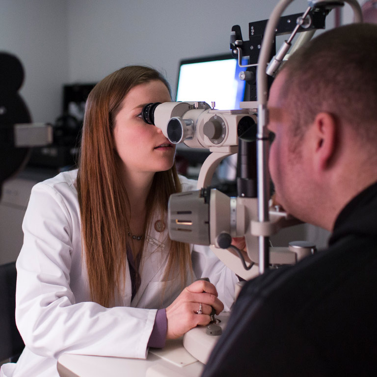 A female optometry student in a white jacket examines a patient's eyes.