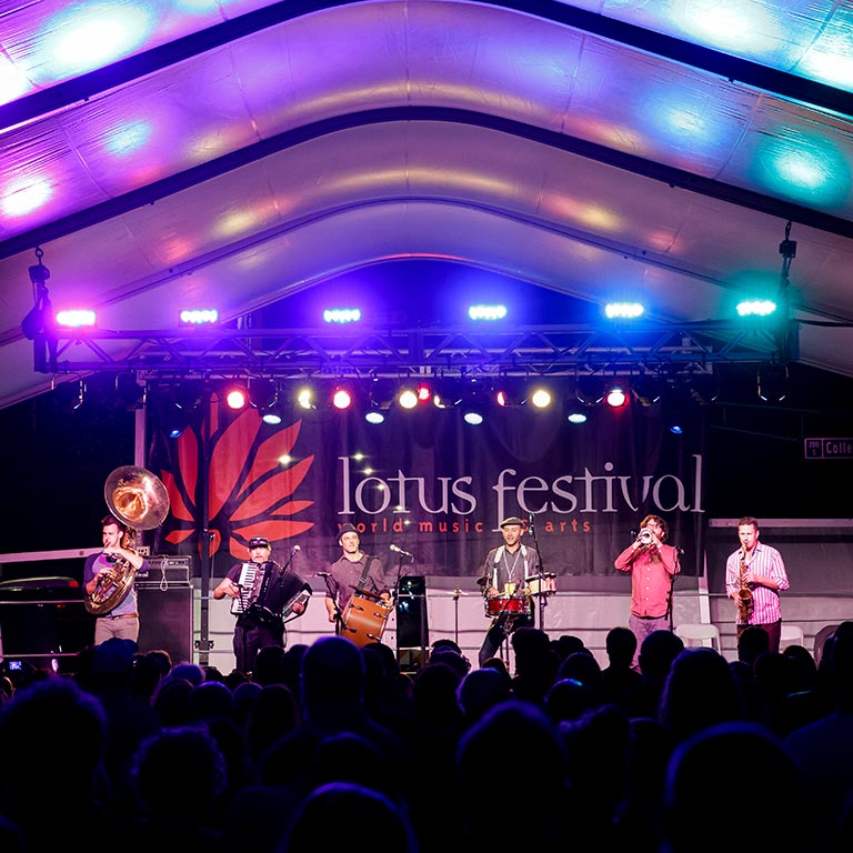 A band with a tuba, accordion, drum, and other instruments play on a brightly lit stage at the Lotus Festival.