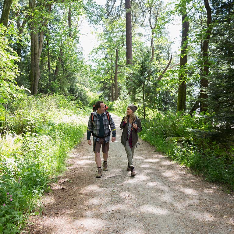 A couple hikes on an outdoor trail in Bloomington, surrounded by tall trees and grasses.