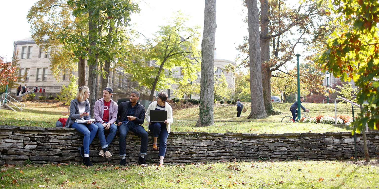 A diverse group of students sit along a stone wall between classes.