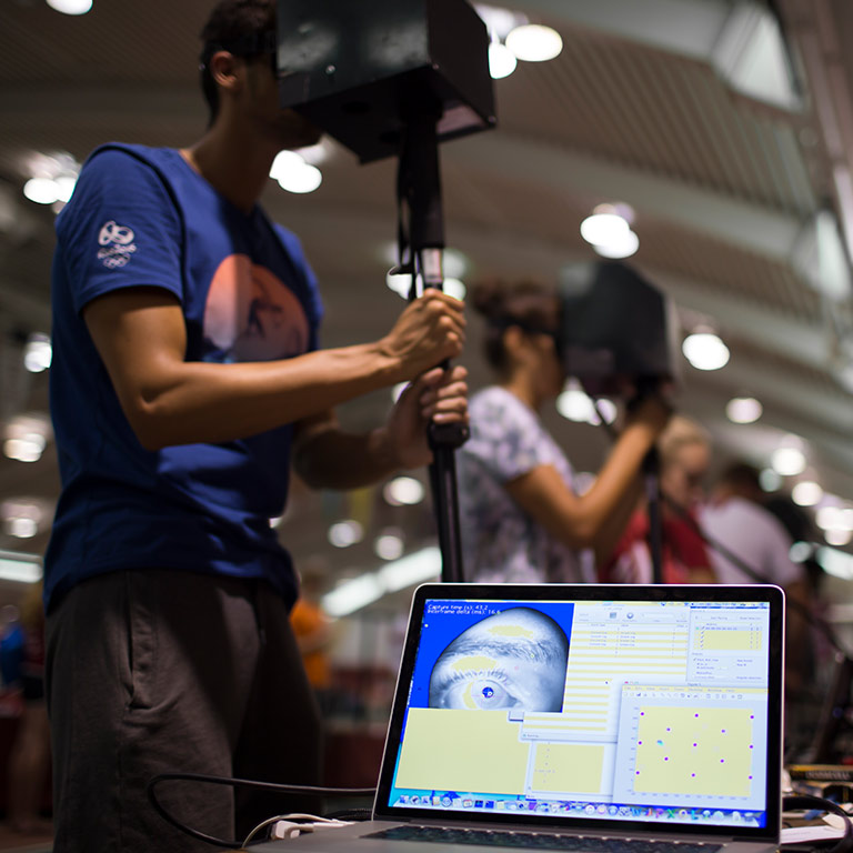 A row of athletes test concussion detection tools developed at the IU School of Optometry. In the foreground, a closeup image of an eye is displayed on a laptop computer.