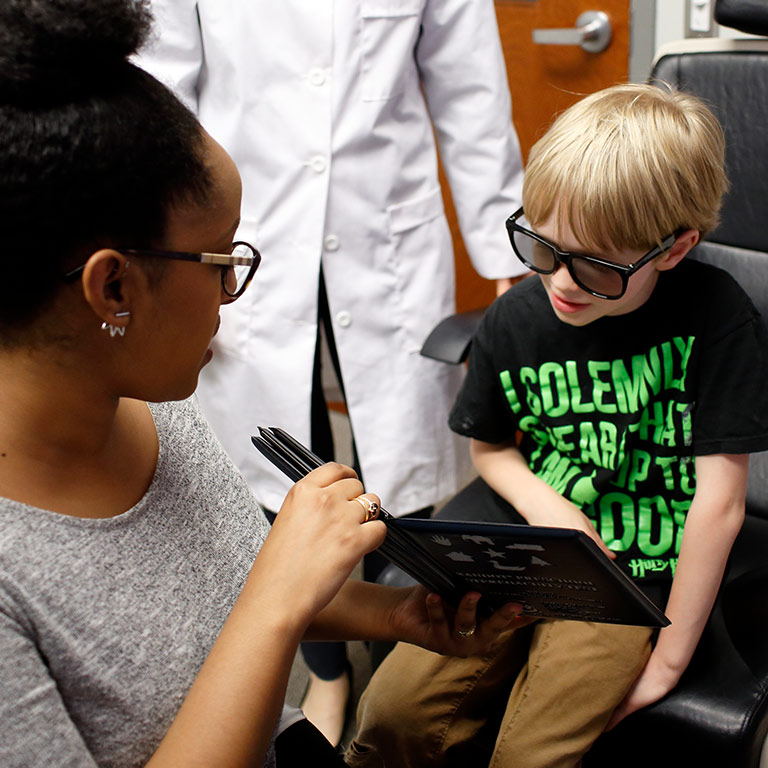 A young boy wears special glasses for his vision exam, while a student holds up a book of shapes. The boy's shirt reads: 'I solemnly swear that I am up to no good. – Harry Potter'
