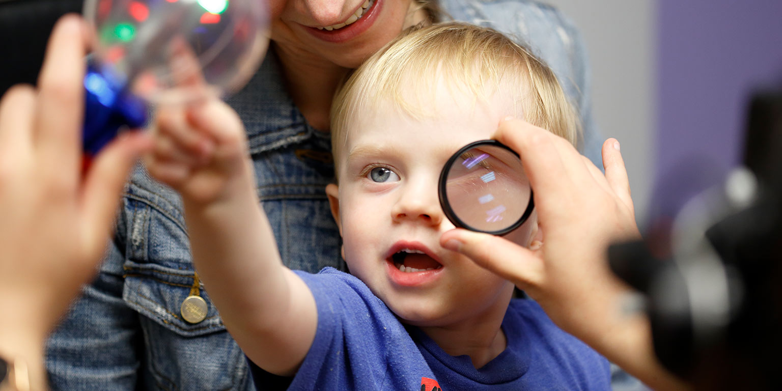 An infant looks at a brightly colored toy while an optometry student examines his eye